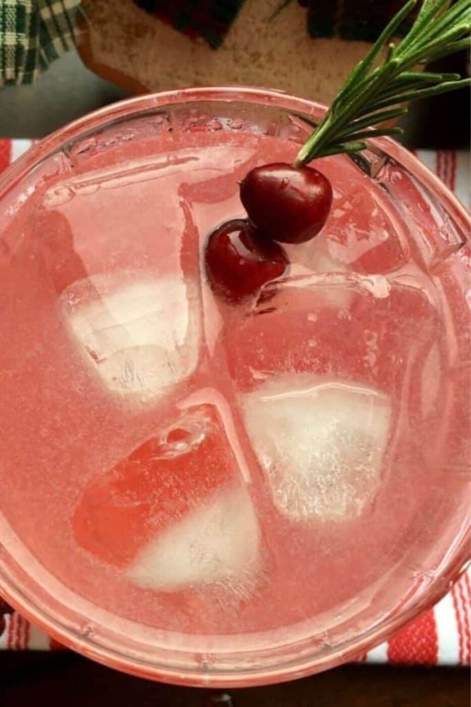 A glass of Cranberry Margaritas on a table decorated with Christmas ornaments. The margarita is garnished with a sprig of rosemary and a fresh cranberry.