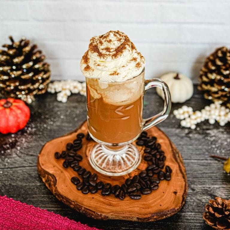 A cup of pumpkin spice coffee with whipped cream on top, sitting on a wooden cutting board surrounded by coffee beans and pumpkins.