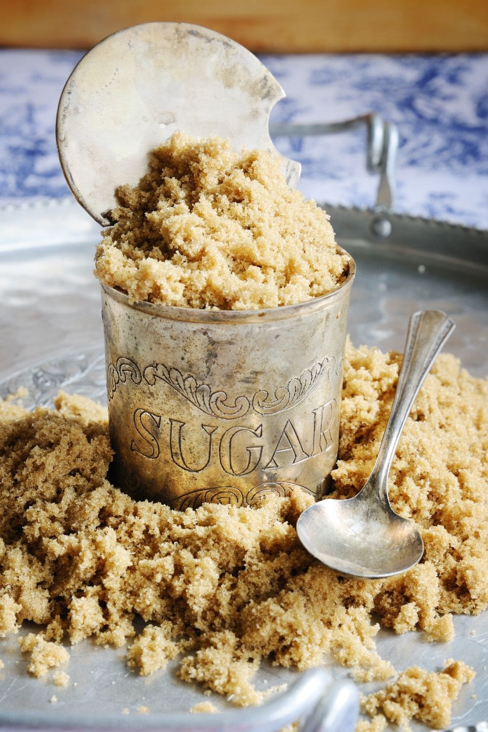 A silver cup filled with brown sugar next to a spoon on a tray filled with brown sugar.