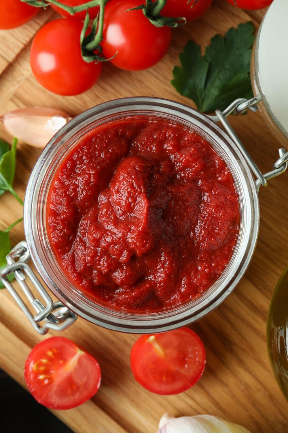 Tomato paste in a glass jar. The jar is sitting on a wooden cutting table surrounded by sliced tomato and basil leaves