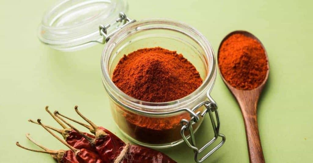 Chili powder in a glass sealable container. It is stilling on a green background and surrounded by chili peppers.