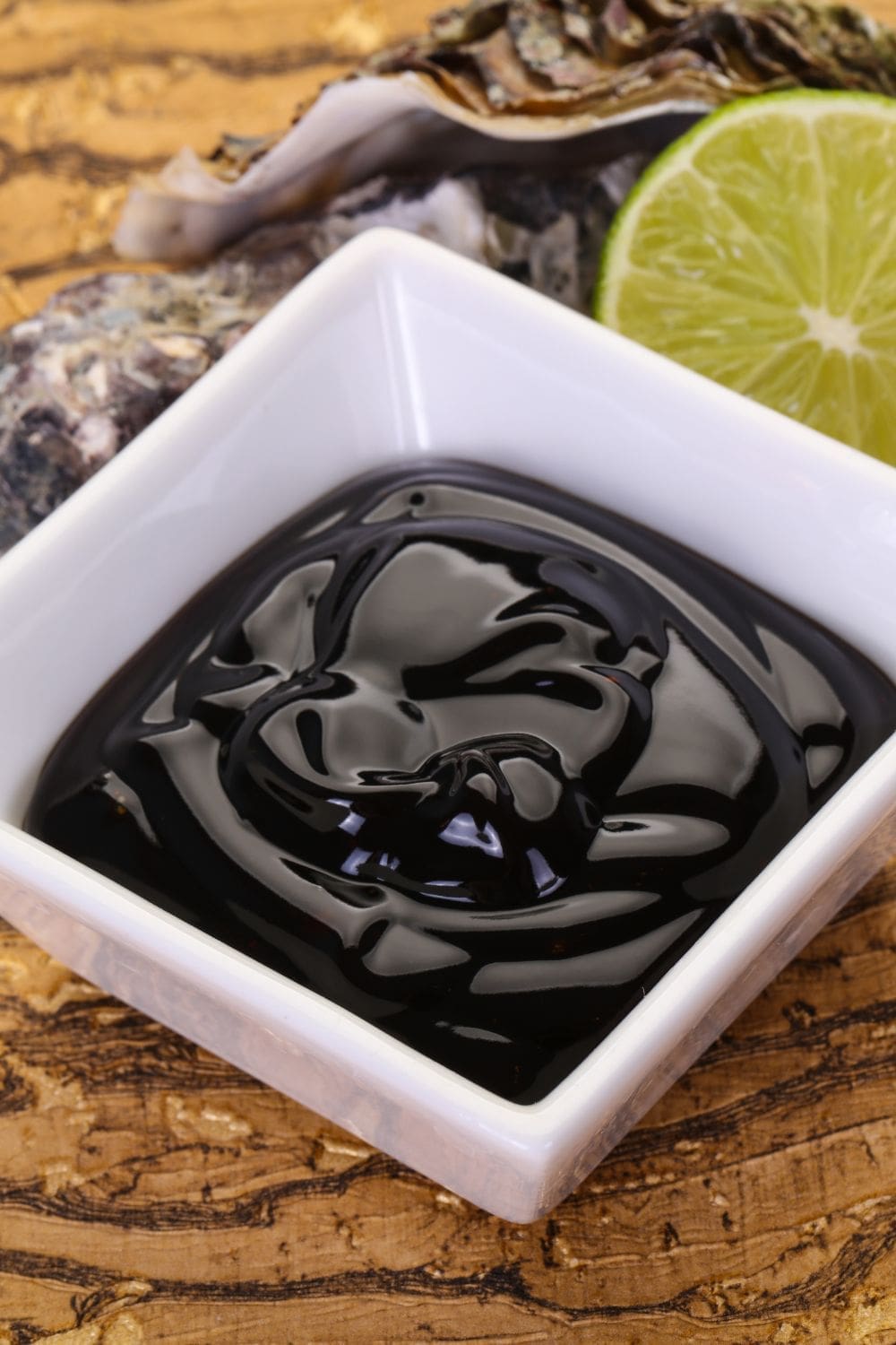 A still life image of a bowl of oyster sauce, an oyster shell, and a lime on a wooden table.