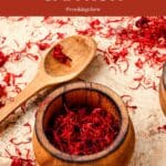 A wooden bowl filled with saffron threads sits next to a wooden spoon on a light-colored table.