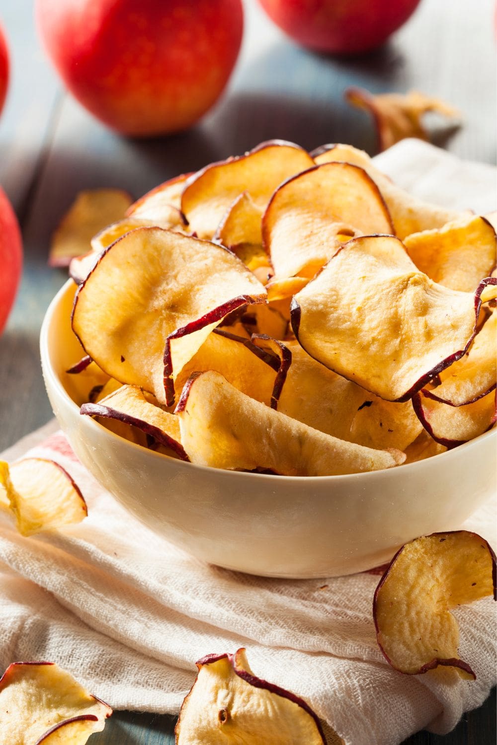 A rustic wooden table with a white bowl overflowing with golden apple chips.