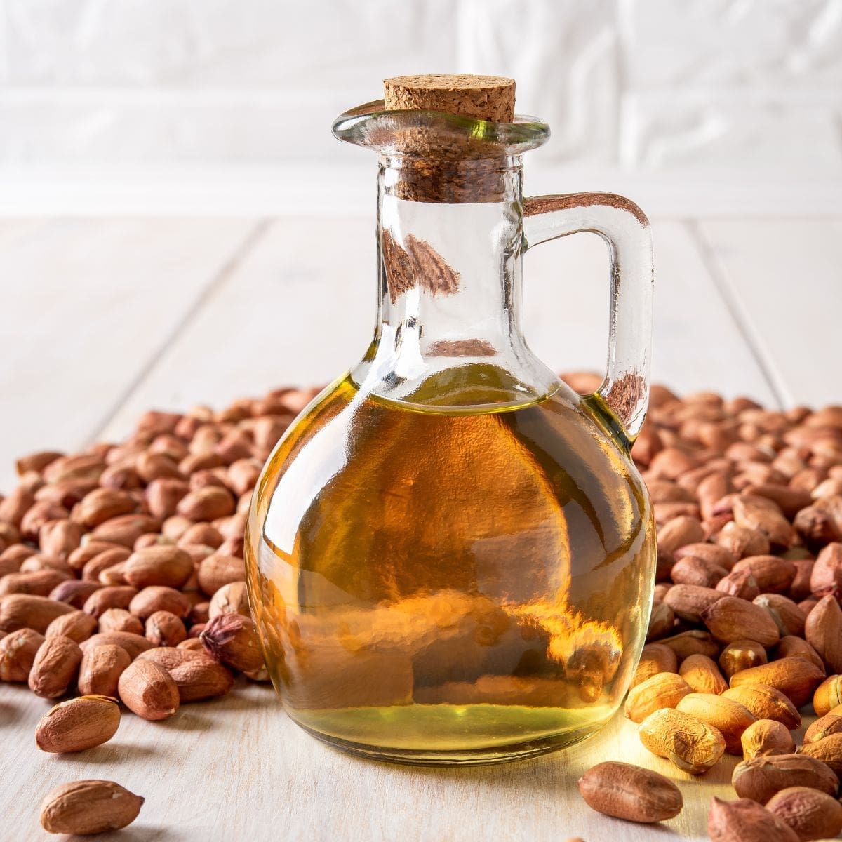 A clear glass pitcher filled with peanut oil sits on a wooden table next to a pile of peanuts in their shells.