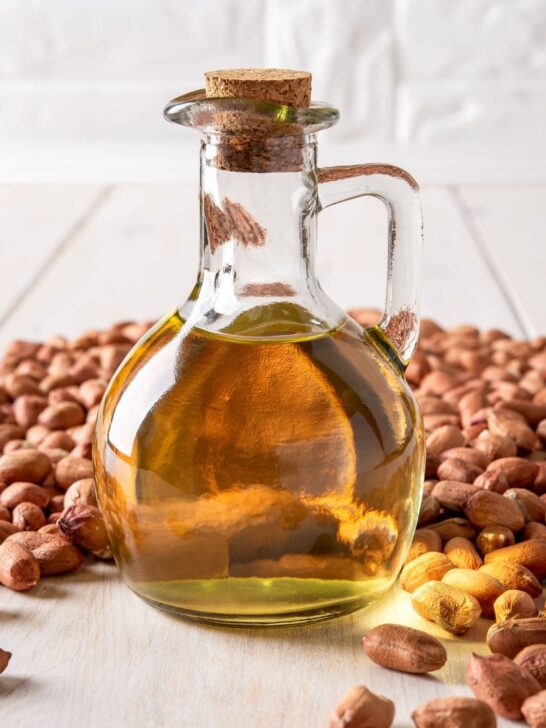 A pitcher of peanut oil surrounded by peanuts on a wooden table.