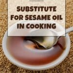 A bottle of sesame oil sits on a wooden table next to a small bowl of sesame seeds.