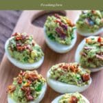 A wooden cutting board topped with deviled eggs filled with avocado and crumbled bacon.