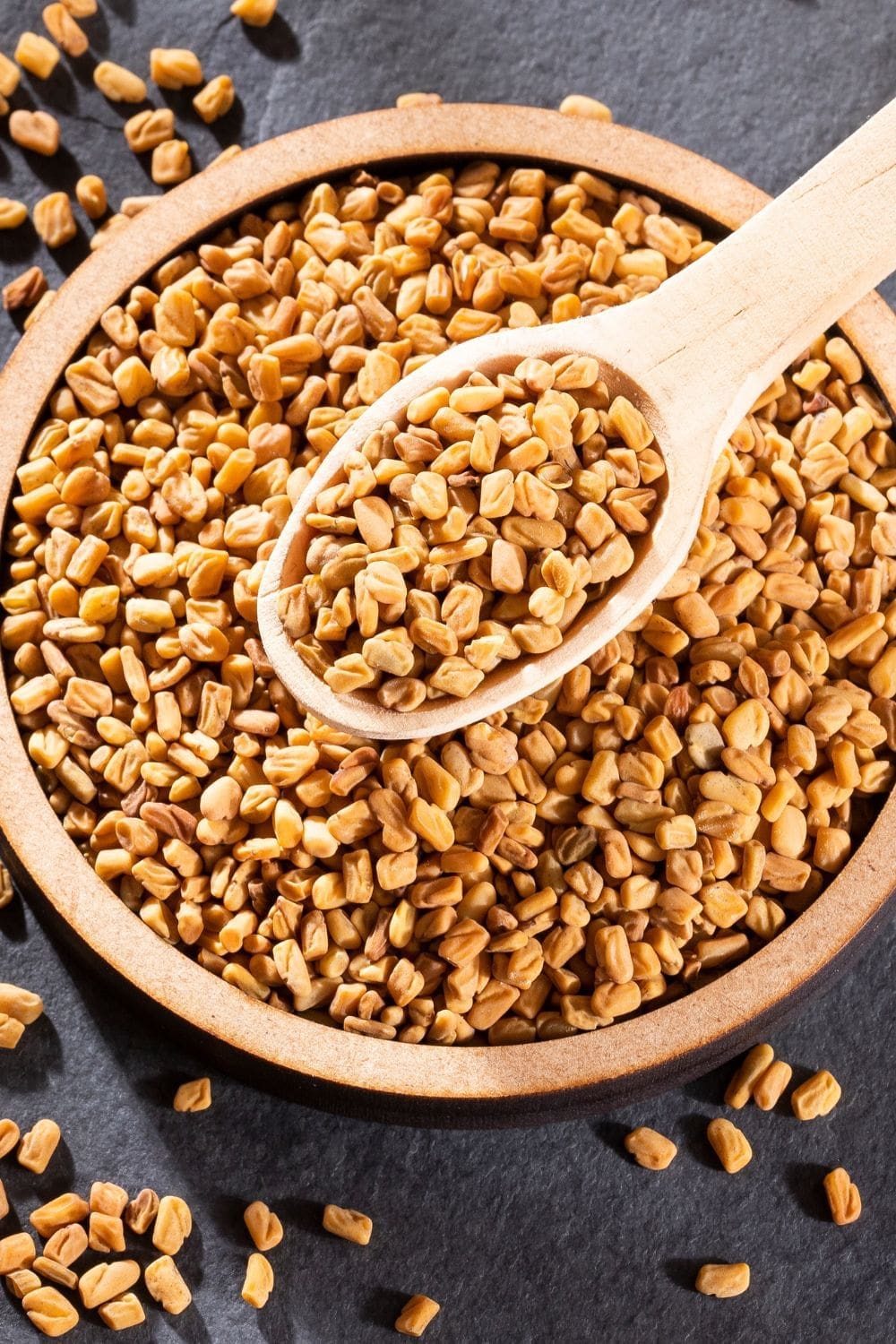 A close-up photo of a rustic wooden bowl overflowing with fenugreek seeds.