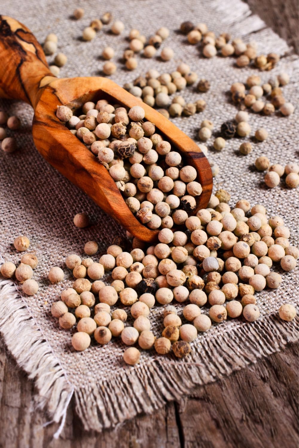A close-up photo of a small wooden scoop filled with white peppercorns. The scoop is on a light-colored wooden table.