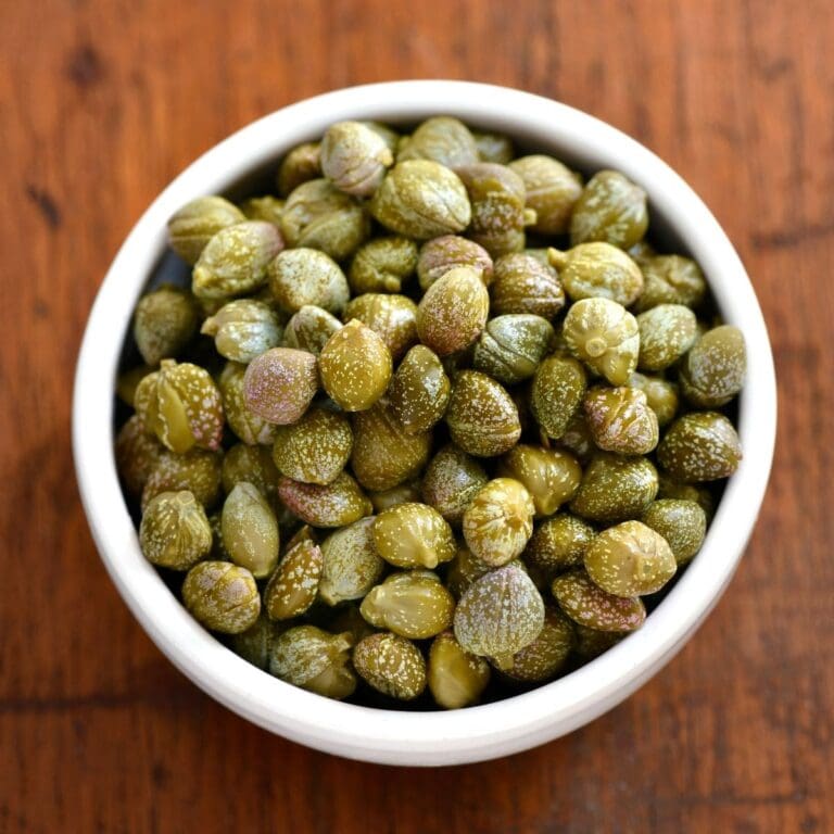 A small, white ceramic bowl filled with green and brown capers rests on a light wooden table.