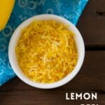 A bowl of lemon peel on a wooden table next to two lemons.