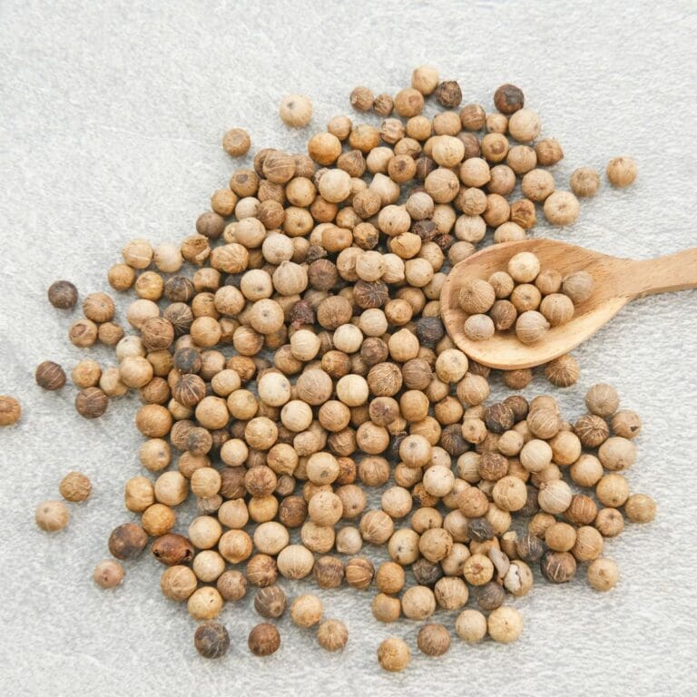 A close-up photo of a pile of white pepper with a wooden spoon on a light-colored wooden table.