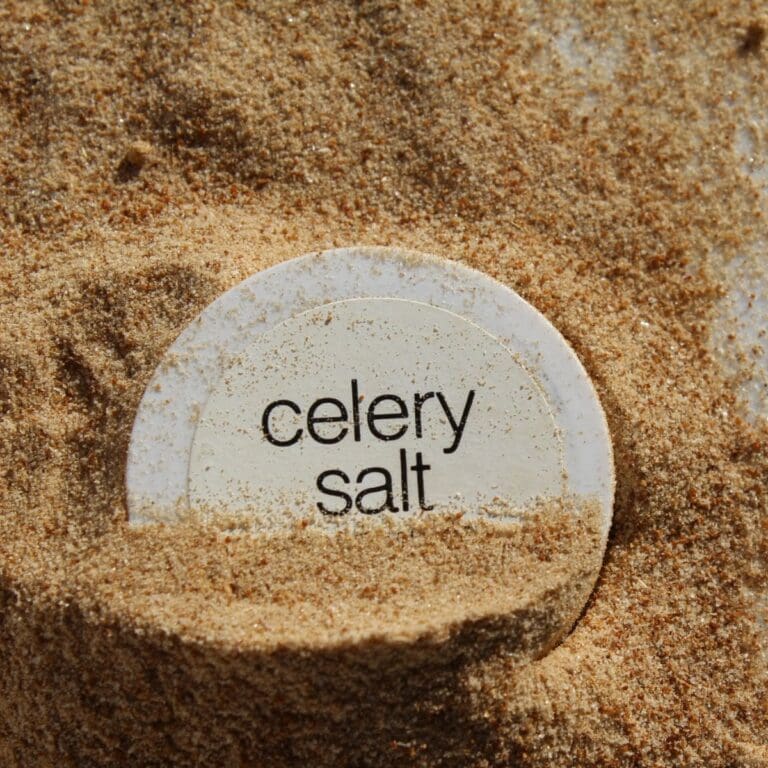 A close-up photo of a round glass jar cap of celery salt next to a small pile of celery salt.