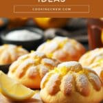 A close-up image of a wooden cutting board piled high with soft, round lemon cookies.