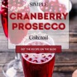 A glass of cranberry prosecco cocktail garnished with a fresh cranberry on a wooden table.