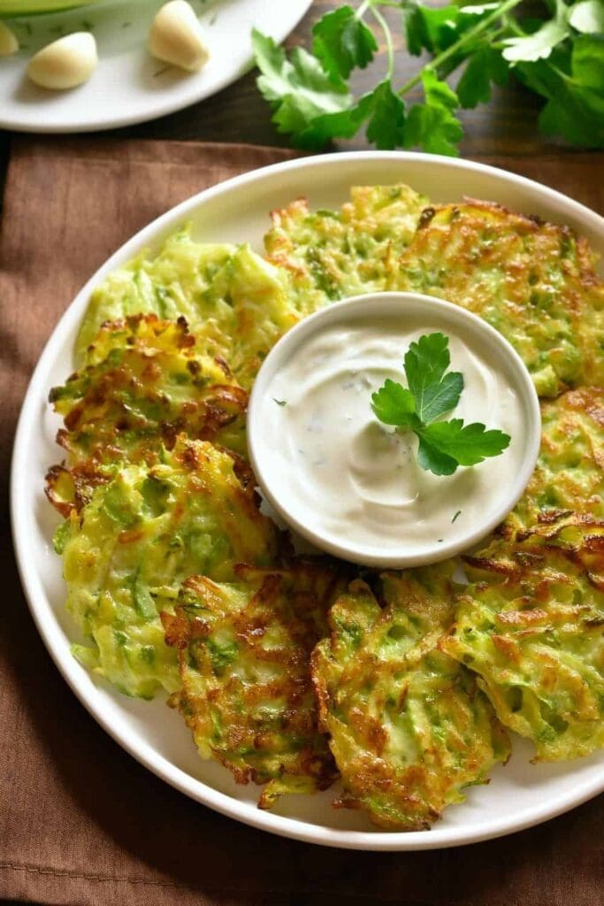 Zucchini fritters spread in a circle around a plate. A dipping sauce is in the middle in a small bowl. 