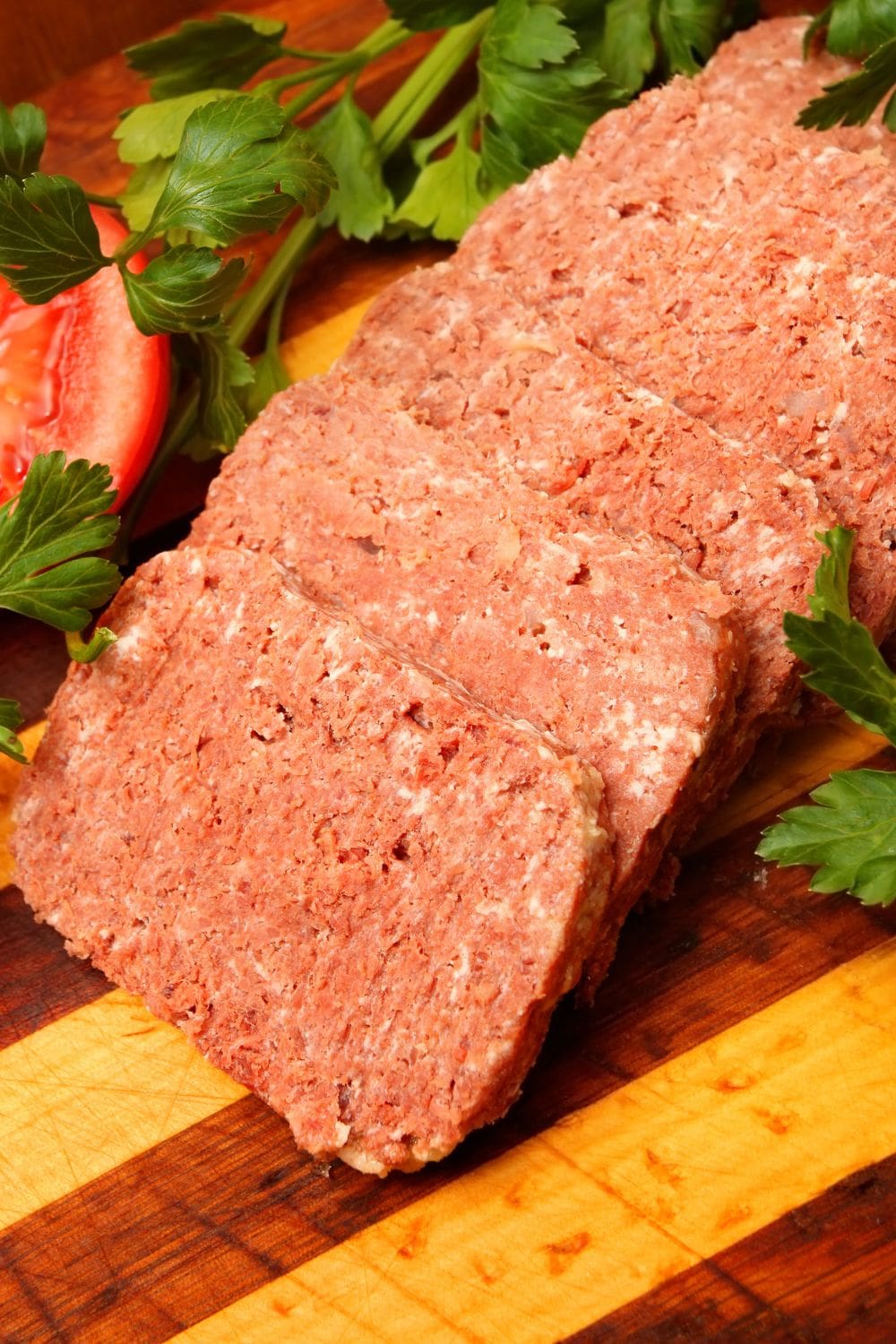 Sliced canned corned beef on a wooden cutting board with a tomato wedge and a sprig of parsley.