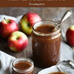 A photo of a glass jar of apple butter next to a pile of red apples on a wooden table.