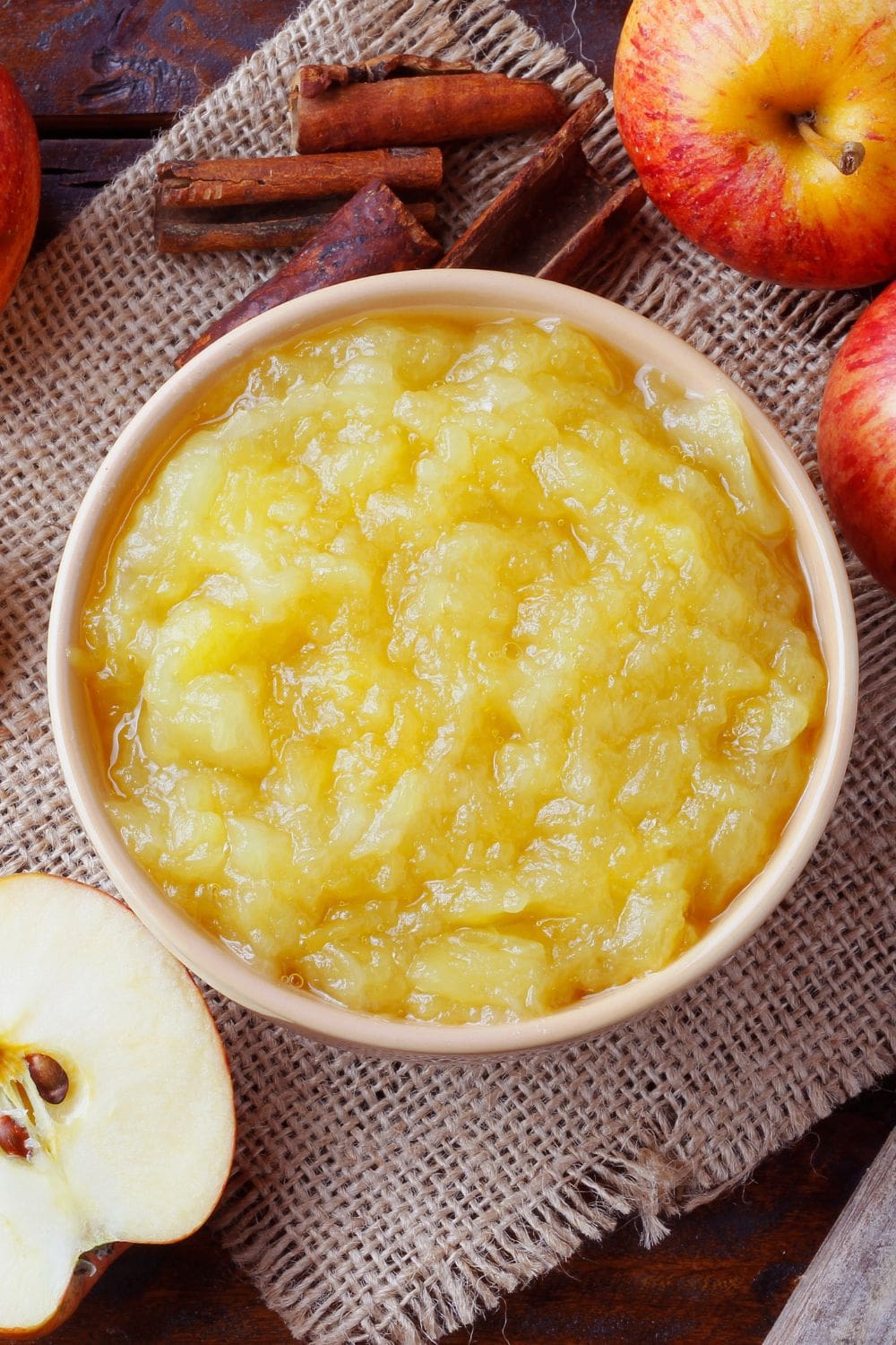 A bowl of apple puree on a table next to apples and cinnamon sticks.