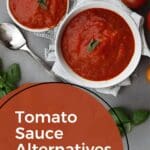 A close-up photo of two bowls of tomato sauce with a spoon on a table with tomatoes in the background.