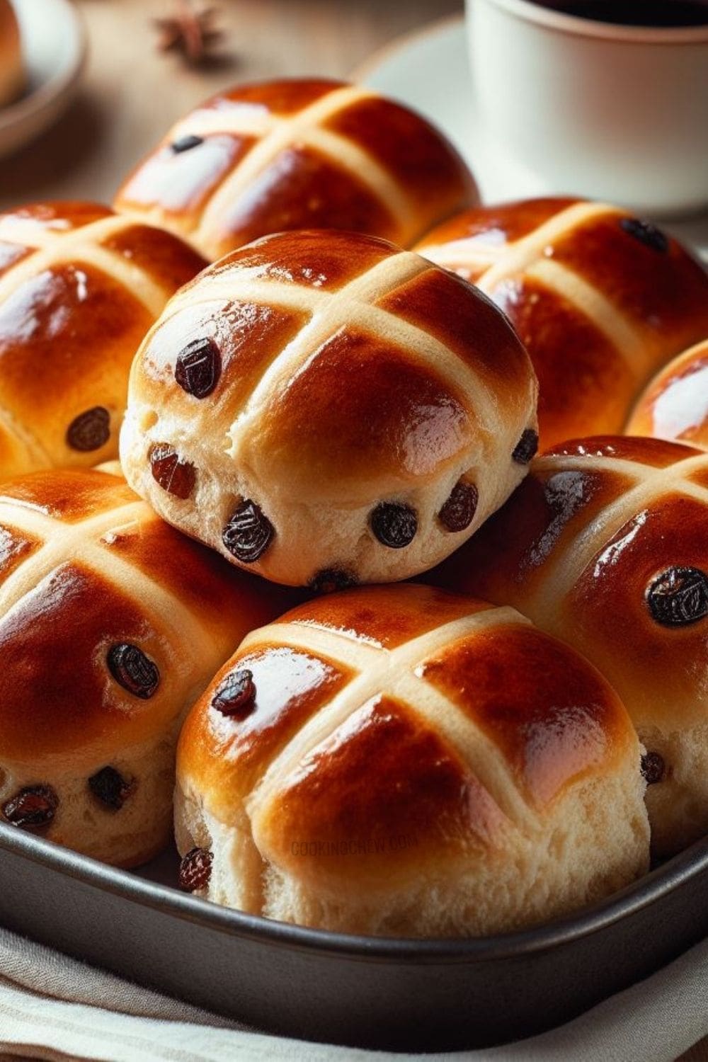 A tray of hot cross buns with raisins, a steaming cup of coffee, and a white napkin on a wooden table.