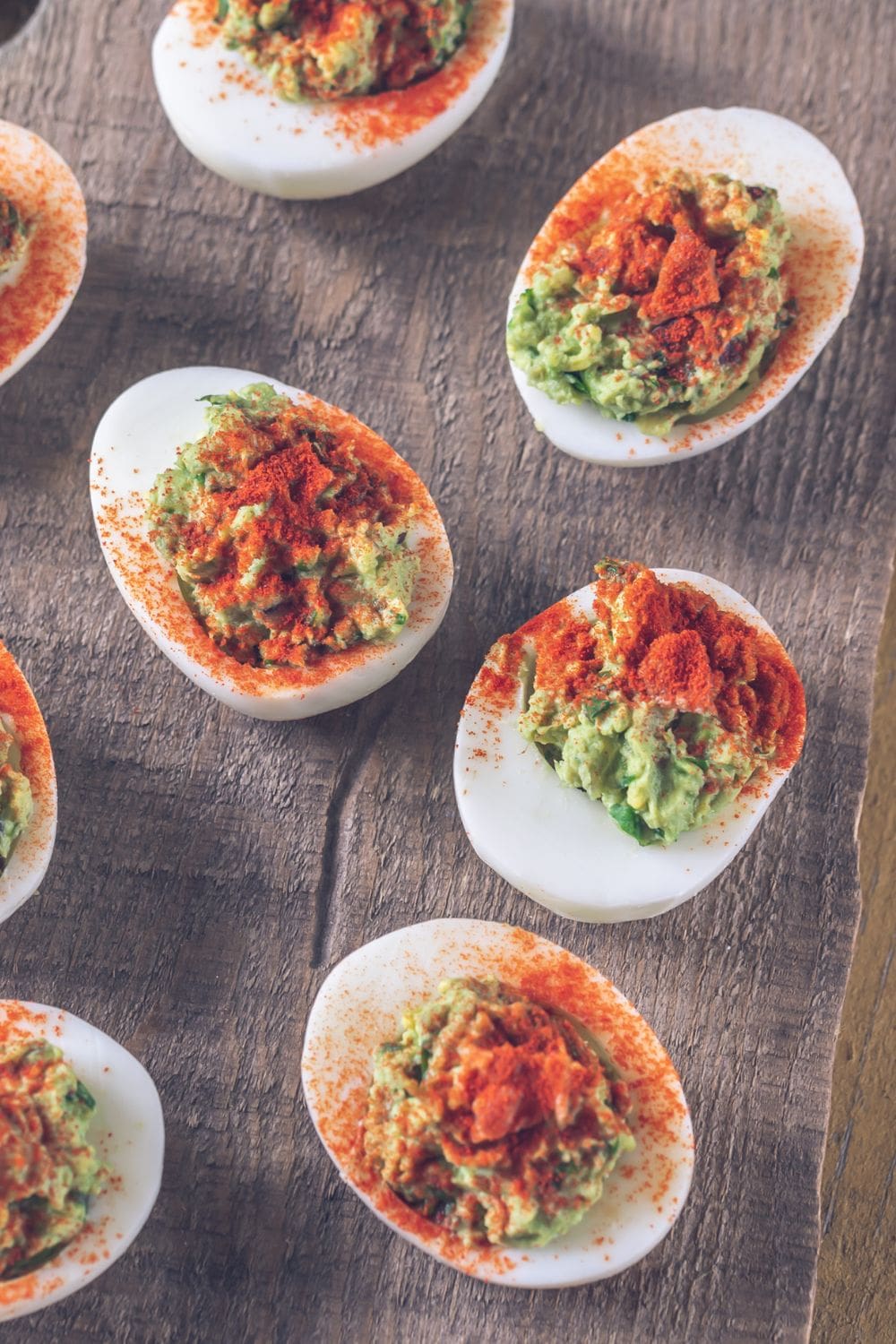 A platter of deviled eggs filled with creamy guacamole and sprinkled with paprika, arranged on a rustic wooden table.