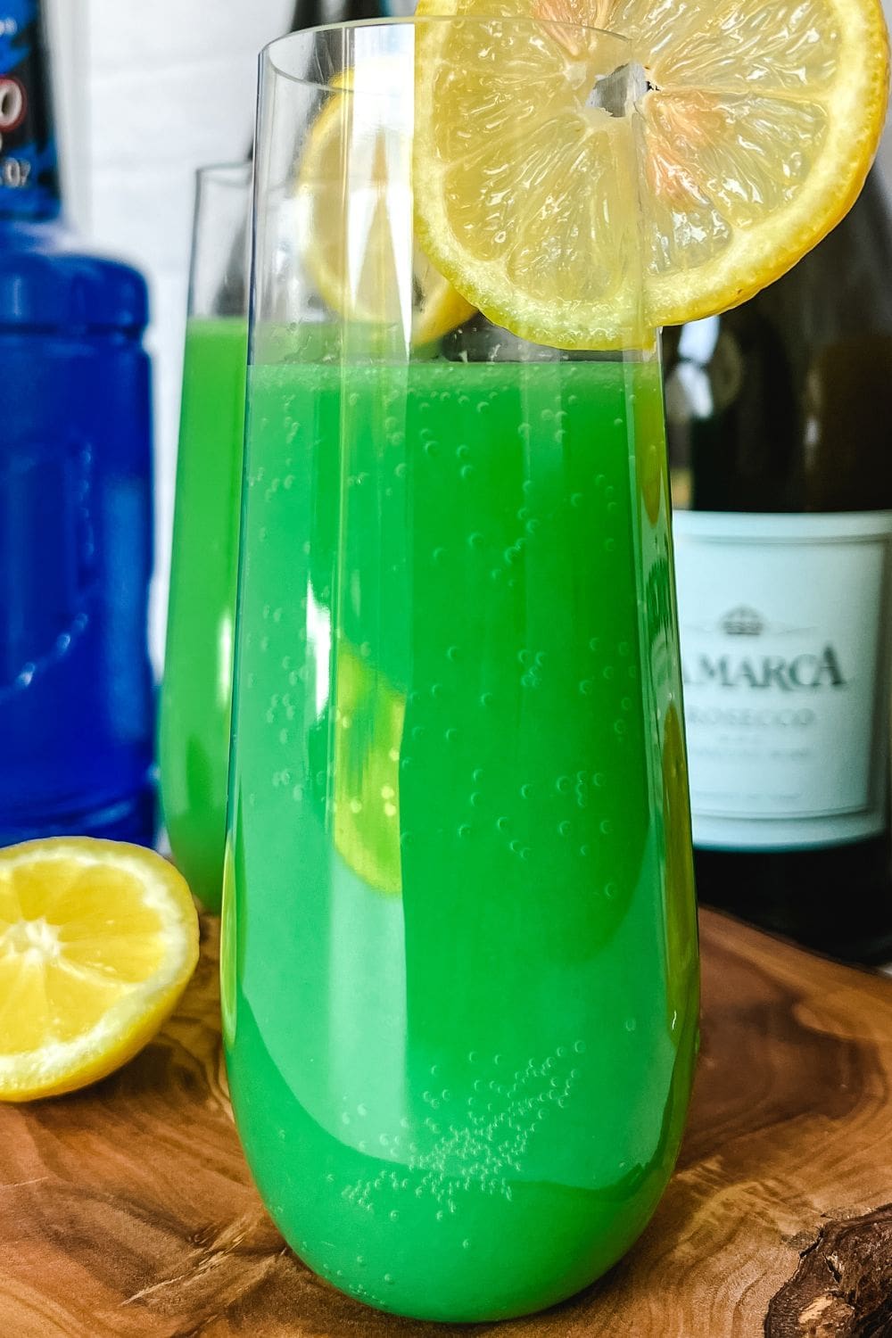 A glass of green beverage with a slice of lemon on the rim sits on a wooden cutting board. 