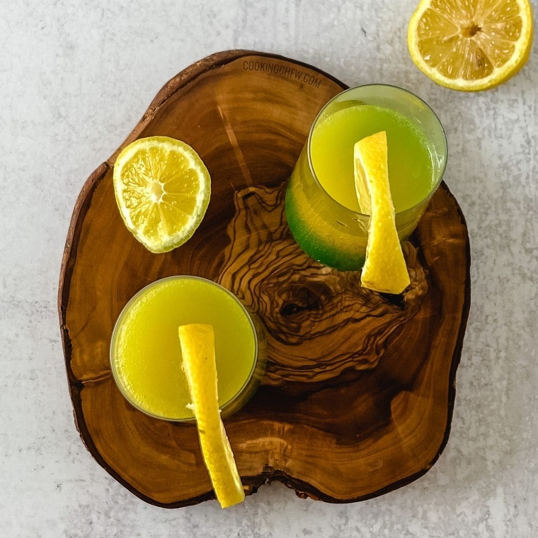 Two glasses of green layered mimosa with ice and lemon slices sit on a wooden cutting board.