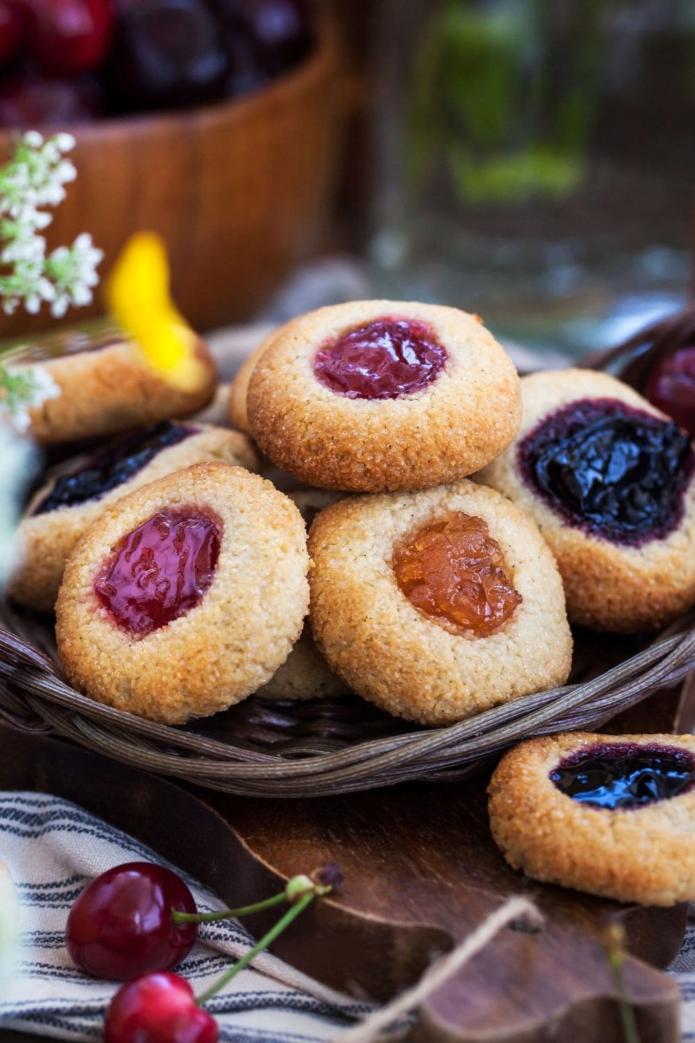 A rustic basket overflowing with homemade jam-filled cookies, accompanied by fresh cherries on a wooden cutting board.