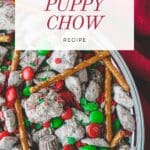 A bowl of Christmas puppy chow, also known as muddy buddies, made with Chex cereal, peanut butter, chocolate, and powdered sugar, topped with Christmas-colored M&Ms.