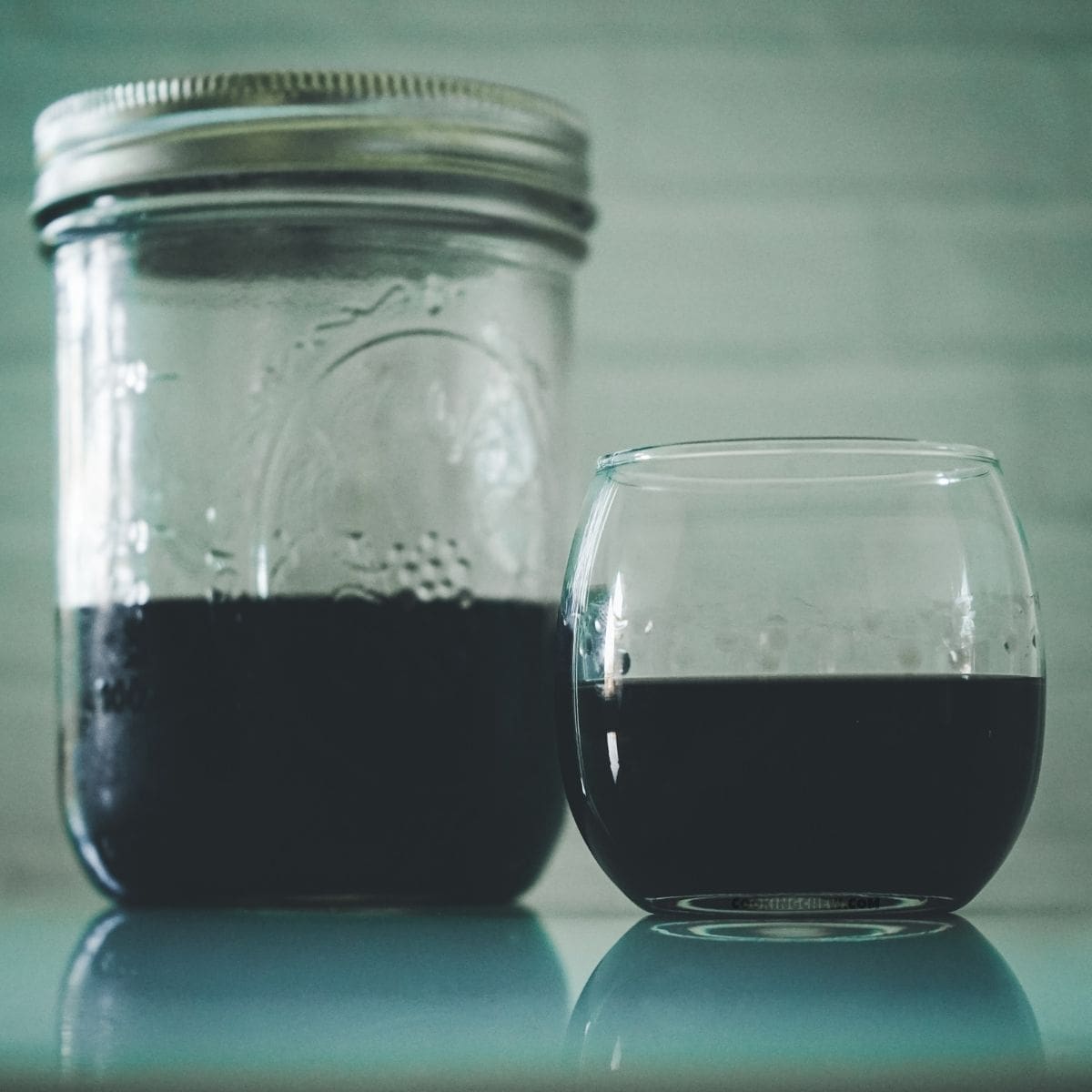 A small mason jar next to a clear glass, both half full with homemade black vodka.