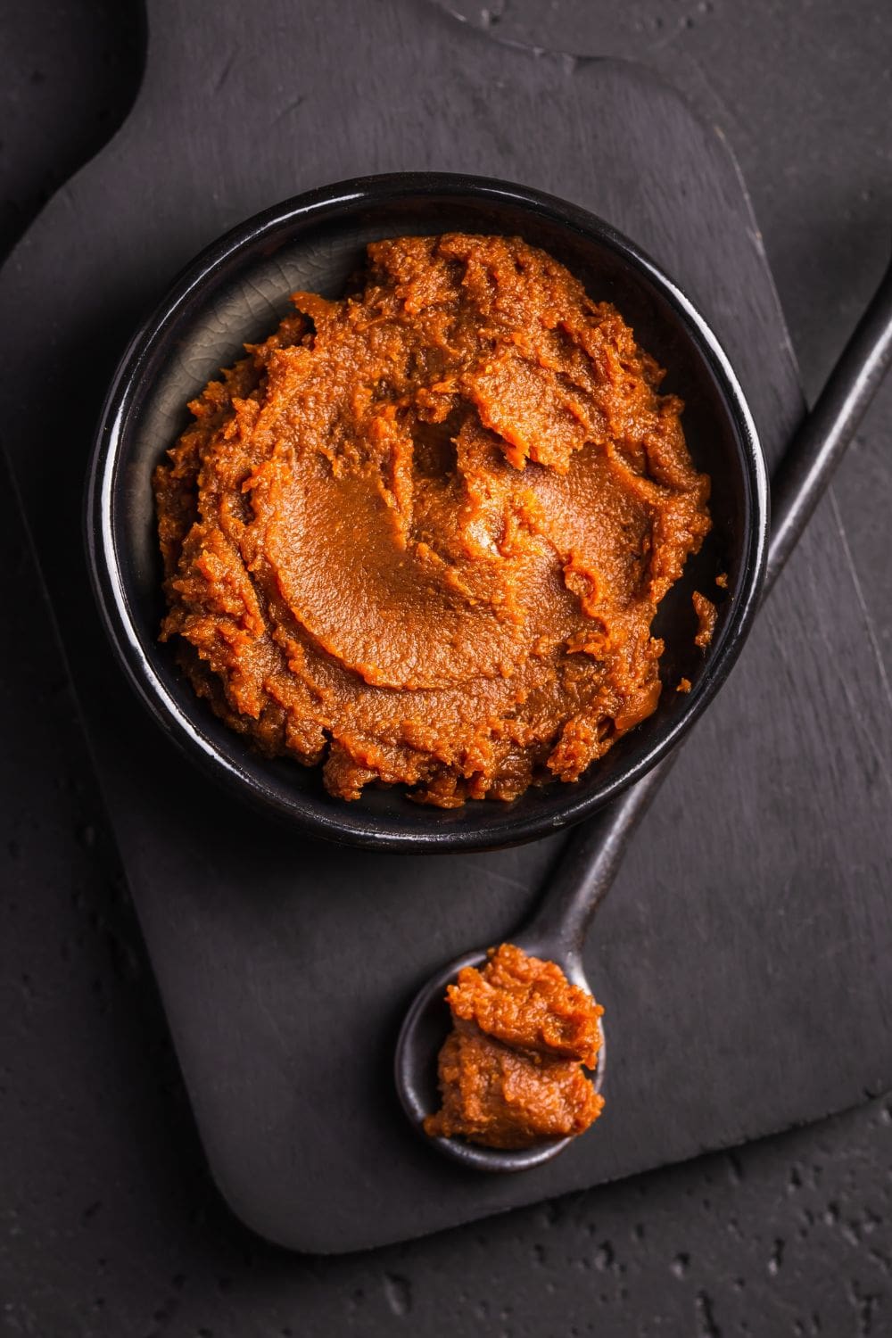 A rustic scene of a bowl of miso paste and a spoon on a wooden cutting board.