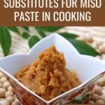 A bowl of thick, brown miso paste surrounded by dry soybeans on a wooden table.