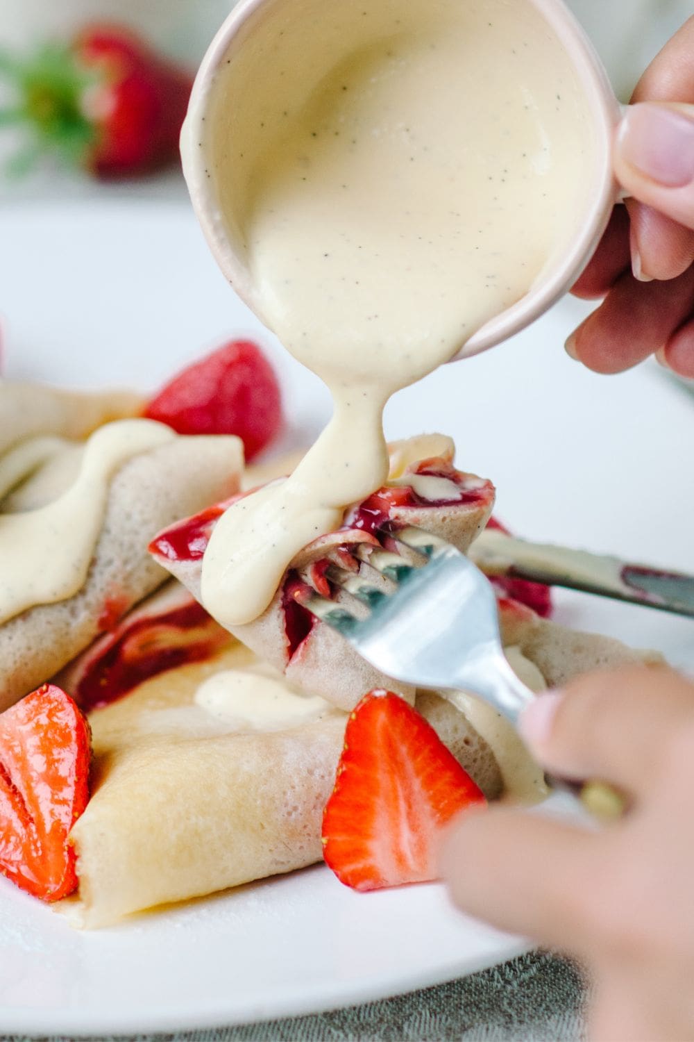 Close-up of a person pouring vanilla sauce from a ladle onto a crepe on a plate.