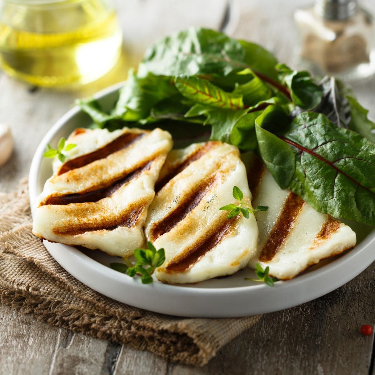 A white plate on a wooden table topped with a halloumi cheese sandwich cut in half and garnished with fresh green herbs.