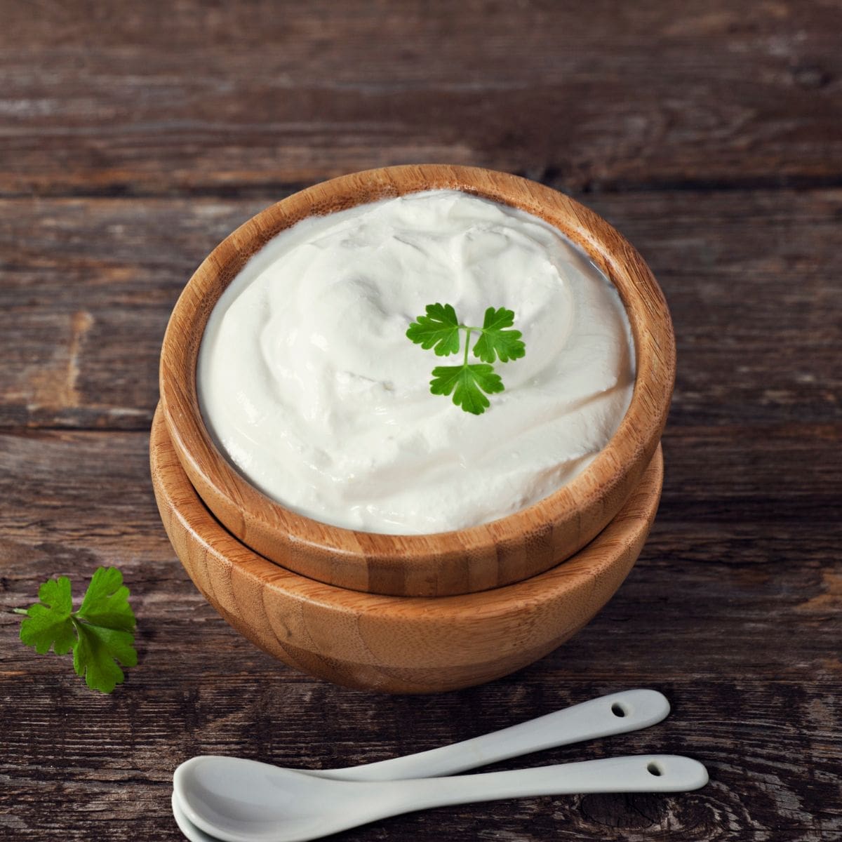 A small white bowl filled with plain sour cream, garnished with a single sprig of curly parsley.
