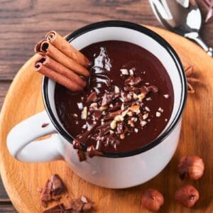 The image shows a cup of hot chocolate with cinnamon sticks and hazelnuts on a wooden table.