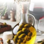 A clear glass bottle filled with green olives sits on a table next to a glass of red wine.