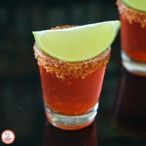 Three vibrant Mexican Candy Shots with Tajín rims and fresh lime slices on a wooden board.