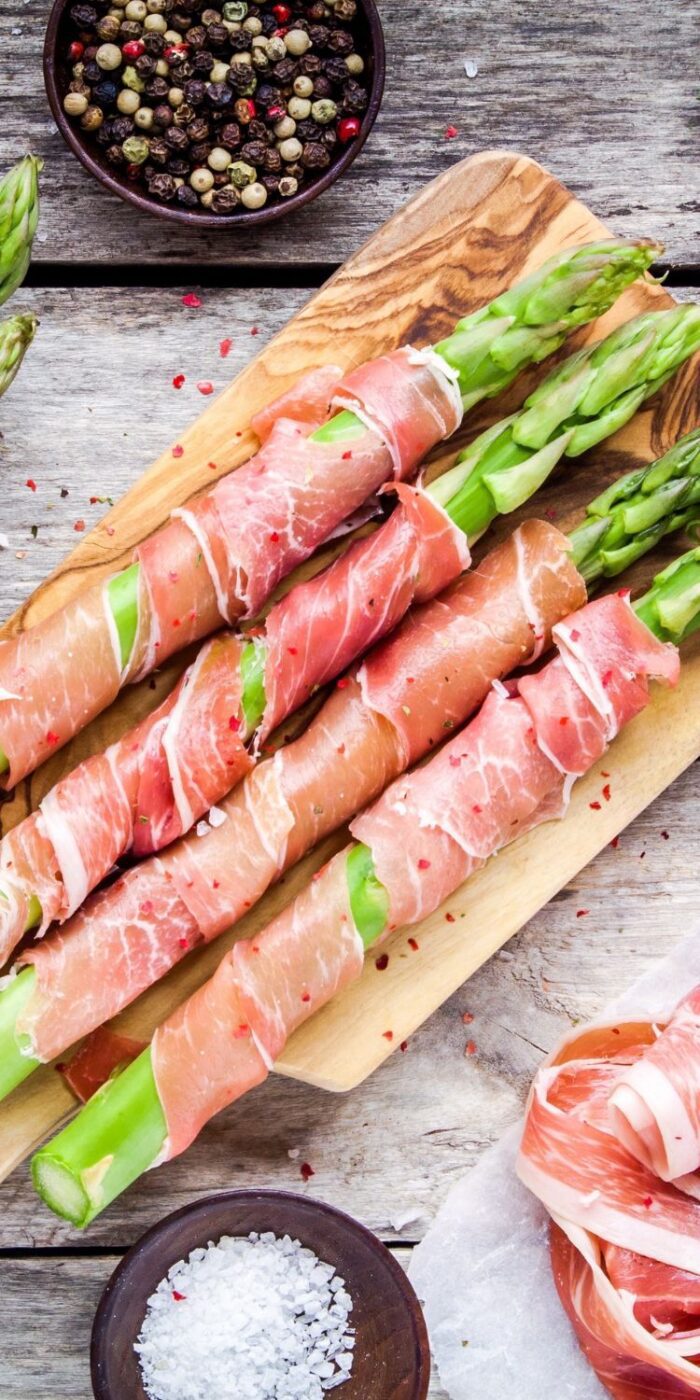 Roasted prosciutto-wrapped asparagus spears on a white plate, perfect for serving with lemon pepper chicken.