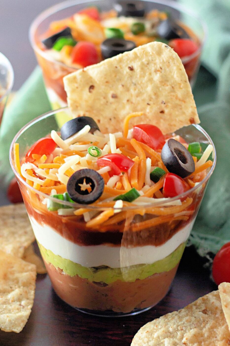 Clear individual cups filled with layers of seven-layer dip, topped with cheese, olives, and tomatoes.