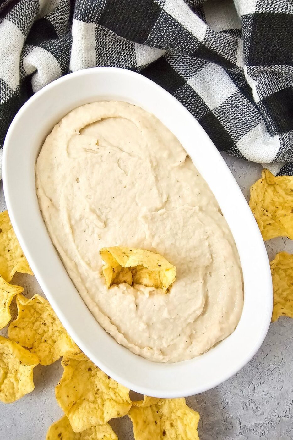 Healthy bean dip in white bowl with checkered towel and chips.
