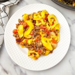 Recipe card photo of cheesy tortellini skillet dinner served with bacon and beef.
