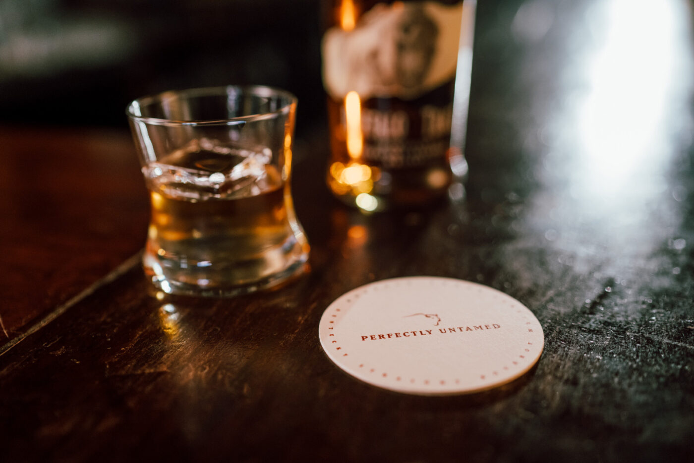 A coaster with the words Perfectly Untamed sits next to a bottle of Bourbon Trace and a glass.