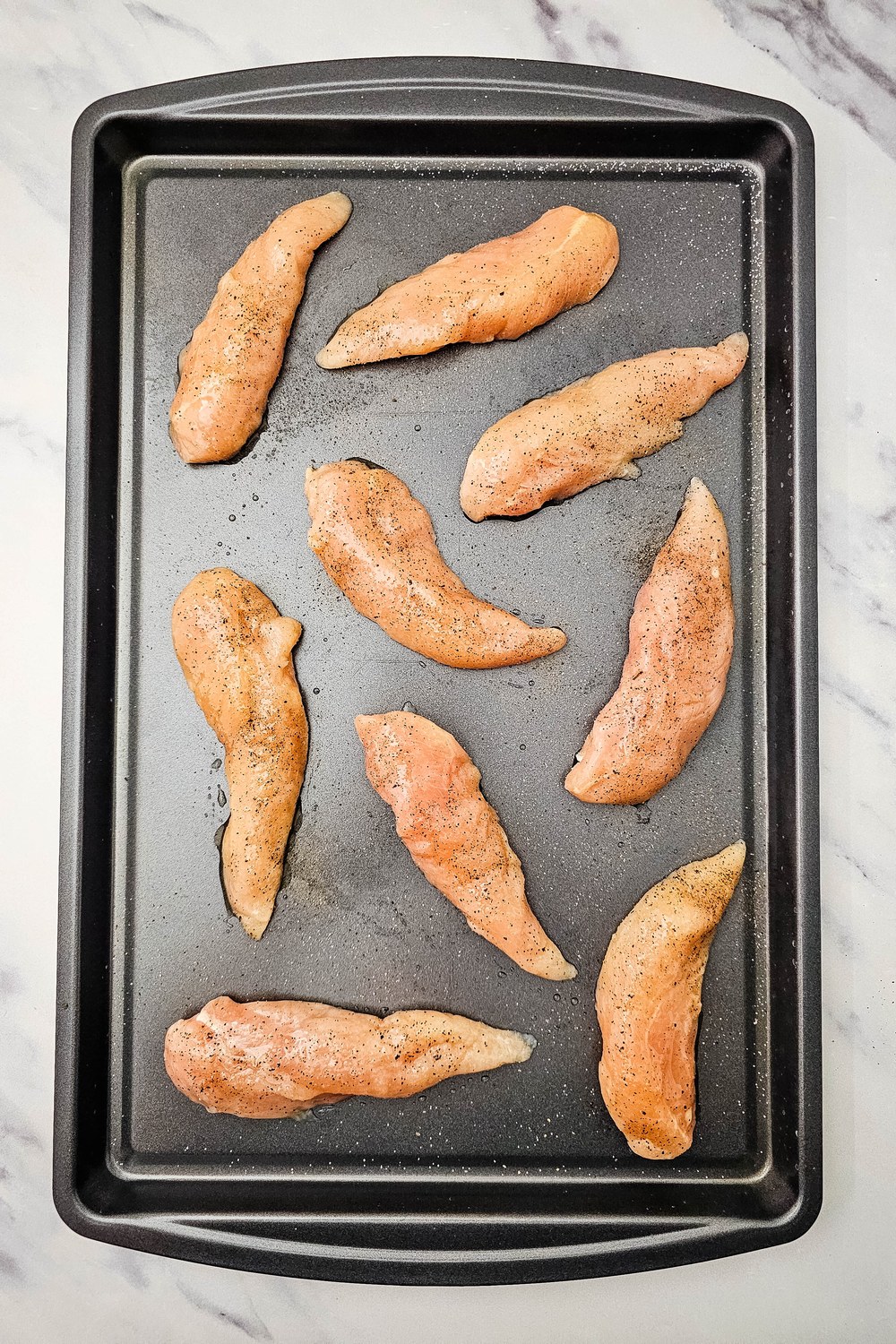 Raw chicken tenderloins arranged on a sheet pan and lightly seasoned with salt and pepper.
