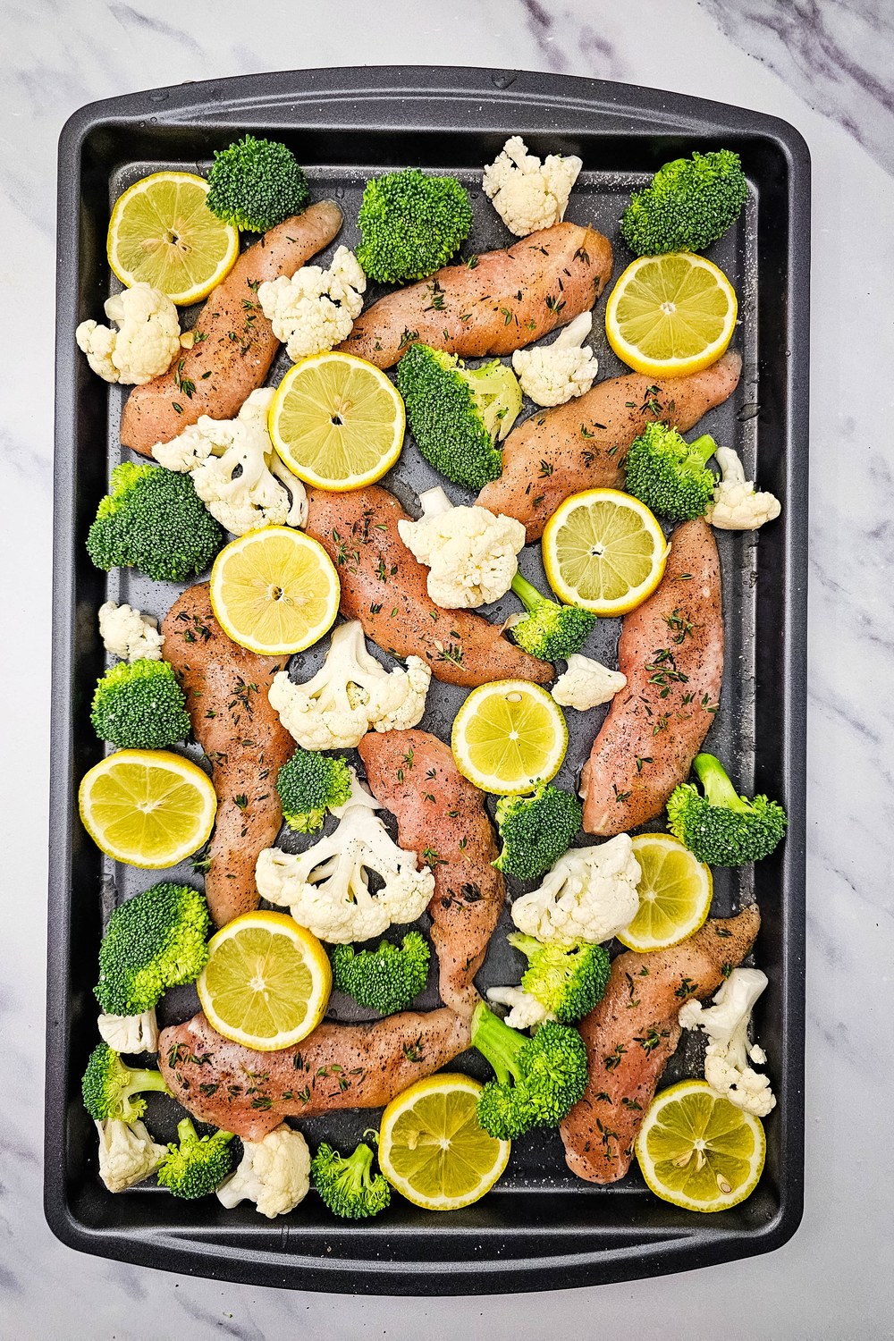 Sheet pan with chicken tenderloins, broccoli, cauliflower, and thin lemon slices ready to bake.