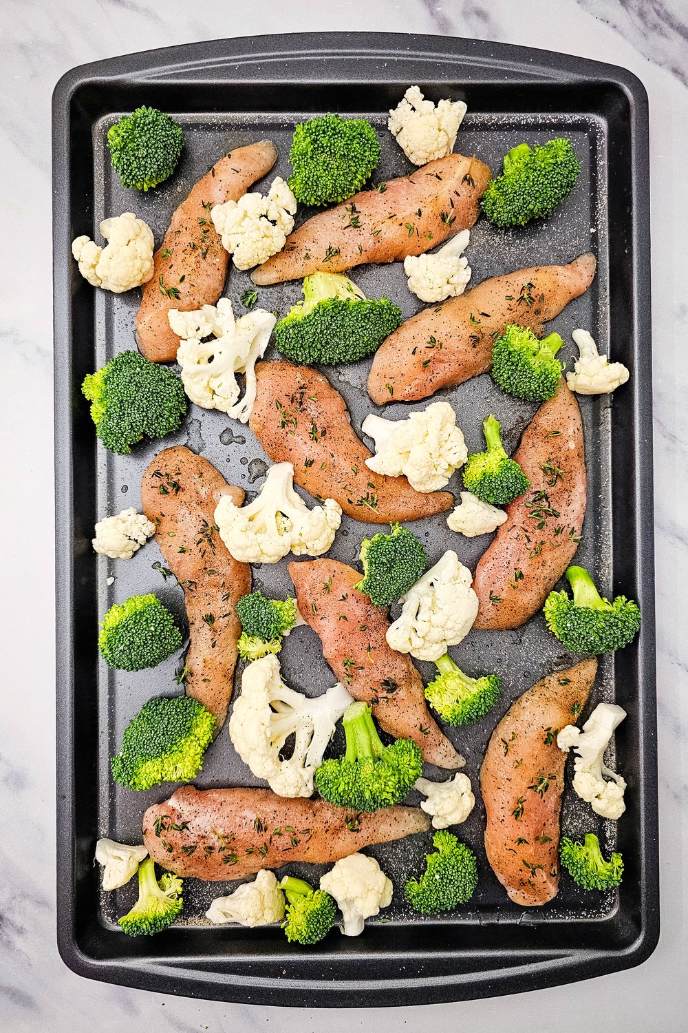 Sheet pan with seasoned chicken tenderloins surrounded by raw broccoli and cauliflower florets.