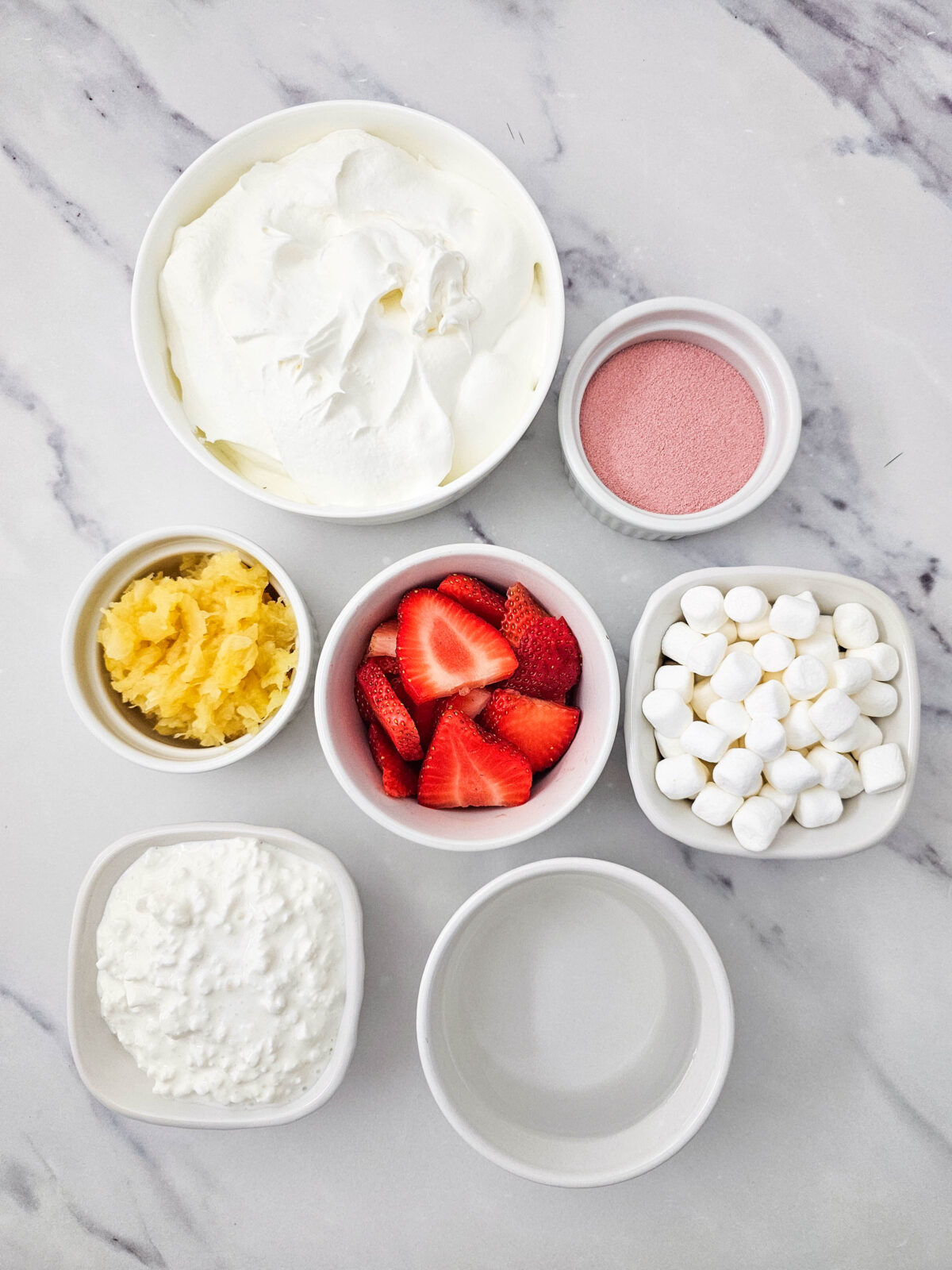 Pink Fluff ingredients laid out on a marble counter: Cool Whip, strawberry Jell-O powder, crushed pineapple, sliced strawberries, mini marshmallows, cottage cheese, and water
