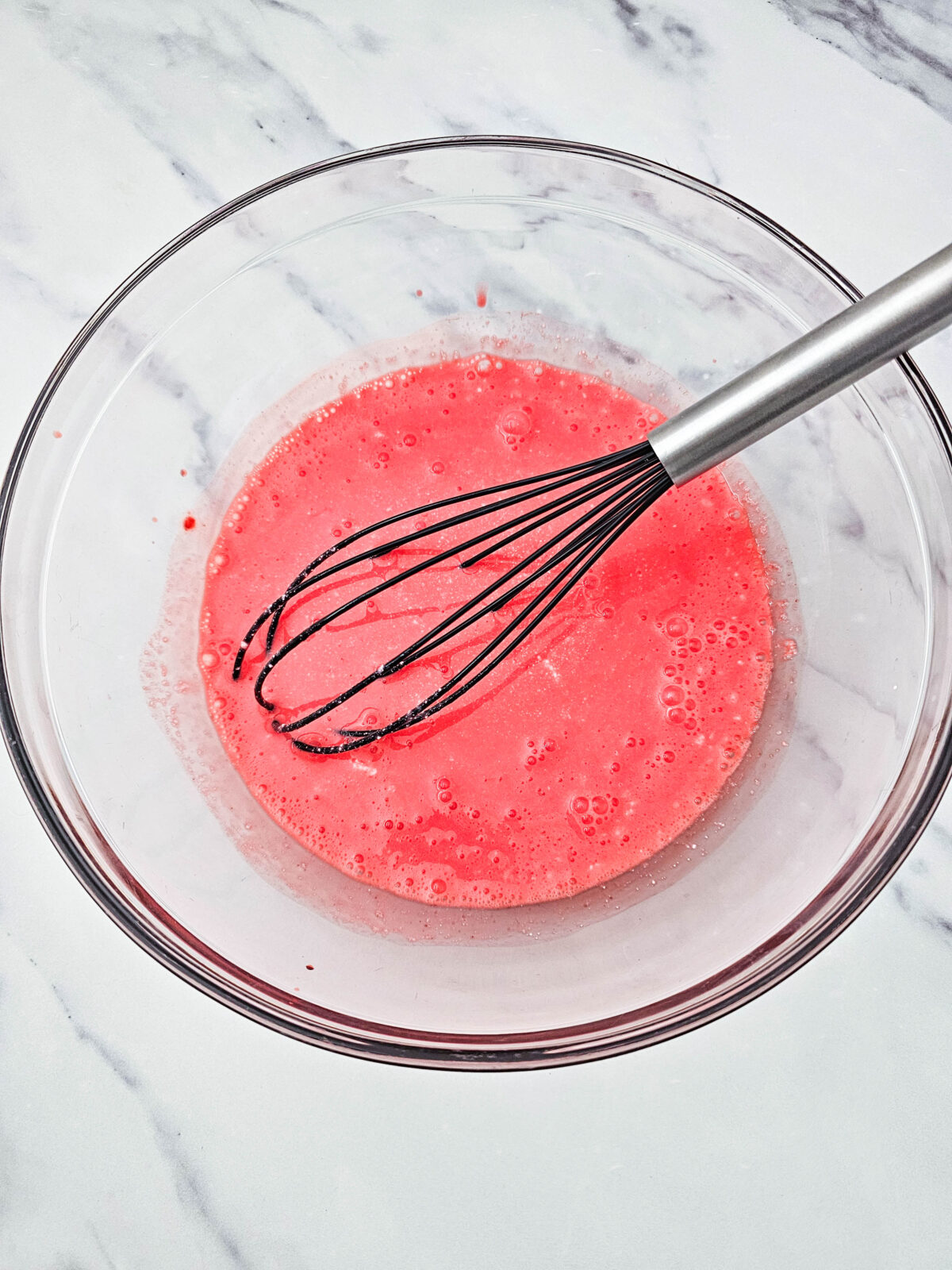 Fully dissolved strawberry gelatin sitting in a glass bowl with a whisk, ready to cool